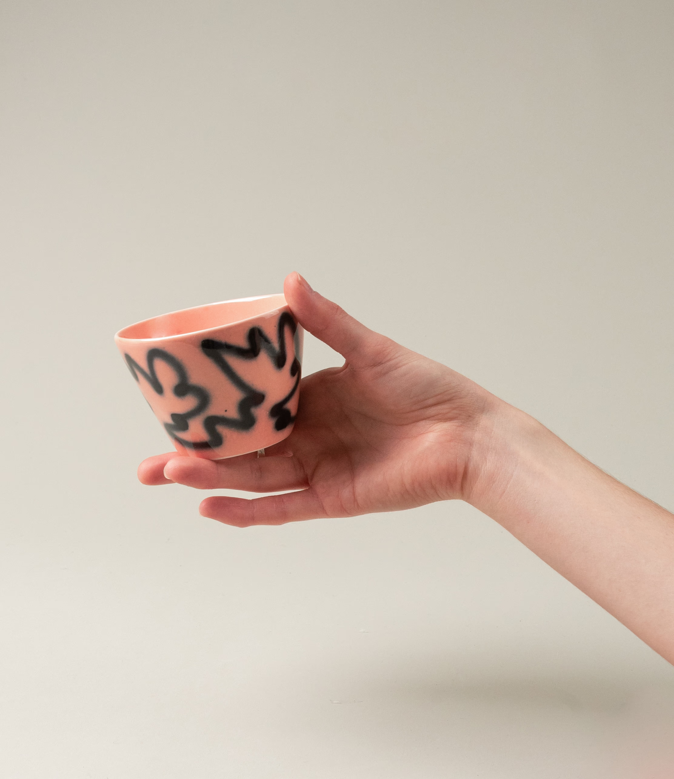 Super Cup from Frizbee Ceramics is a set of two cups. The cups' base is peach with a black graffiti like zig-zag painting on it. On this picture a hand holds the cup.