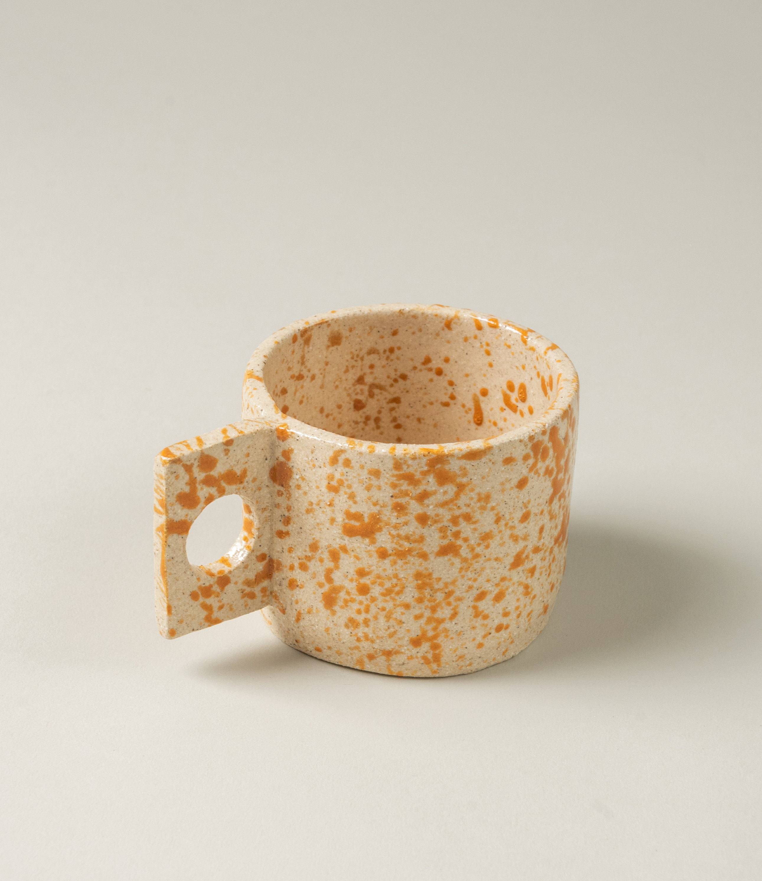 Pumpkin Spice Coffee Cup from Manamo Cups. The product has a natural touch and a modern shape with the rectangular handle. The item has a spotted orange texture on a beige base.