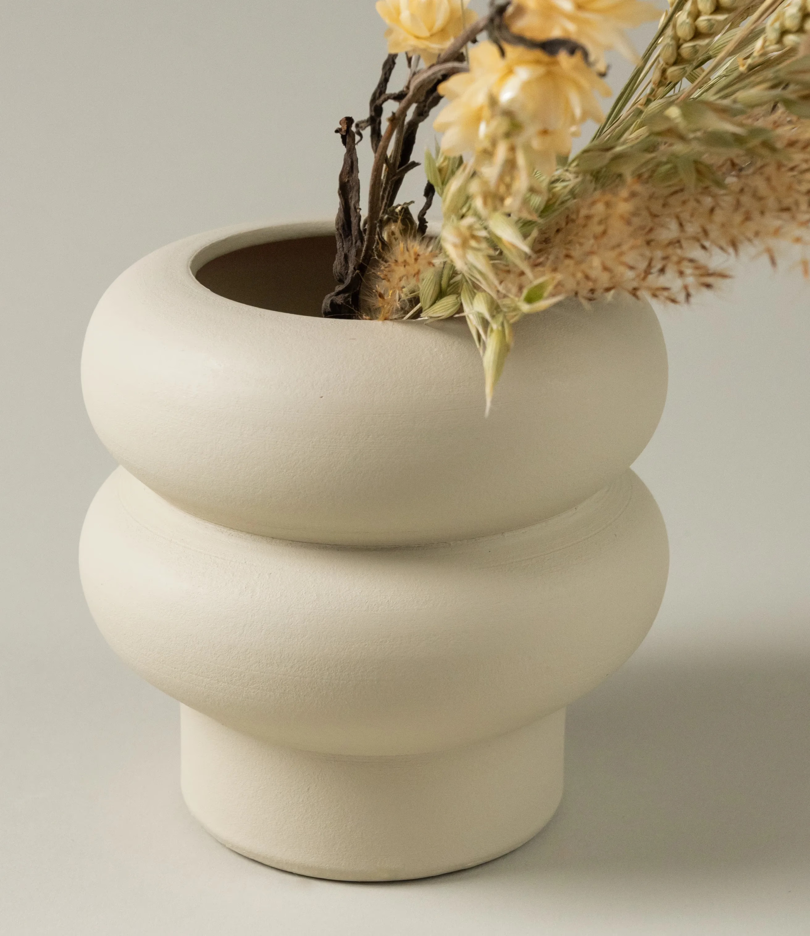 Ocactuu's Ripple Vase coming in the light sand color, and in use with our Natural Dried Flowers in the natural color. The Vase have a curved shape, looks like two rings stacked on top of each other.