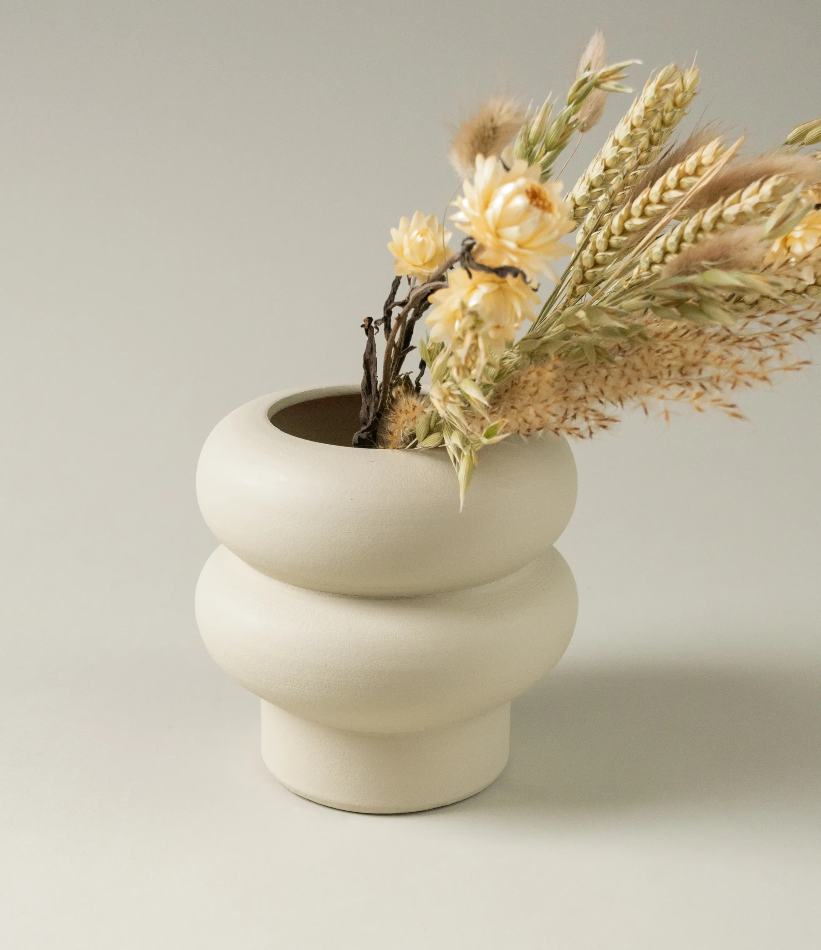 Ocactuu's Ripple Vase coming in the light sand color, and in use with our Natural Dried Flowers in the natural color. The Vase have a curved shape, looks like two rings stacked on top of each other.