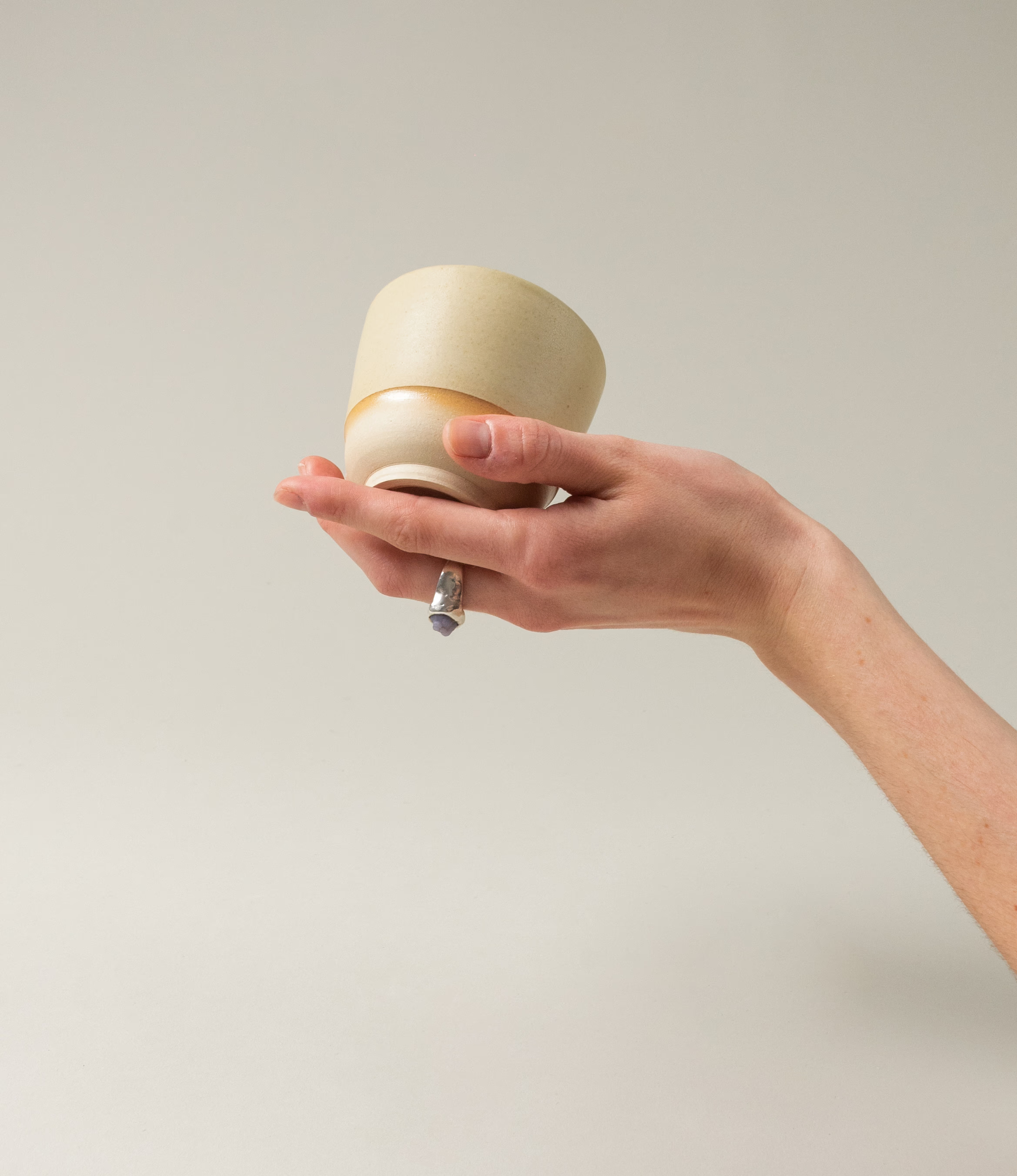 This cup set is handcrafted by Omni Ceramics. The two small cups are wearing a gradient orange color ring on the natural beige base. In this picture the cup is hold by a hand.